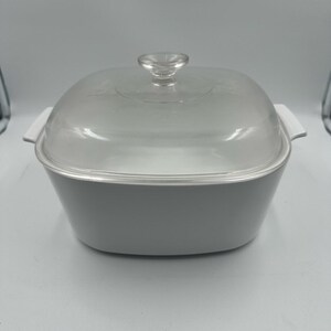 Puede incluir: Una fuente cuadrada de color gris claro con tapa de cristal transparente y pomo transparente. La fuente tiene asas blancas a cada lado. La tapa es ligeramente abovedada. La fuente está sobre una superficie blanca.