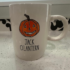 May include: White ceramic mug featuring a hand-drawn orange Jack O'Lantern design with a smiling face and a black stem. The words "JACK O'LANTERN" are written below the pumpkin in black lettering. The mug has a large, curved handle.