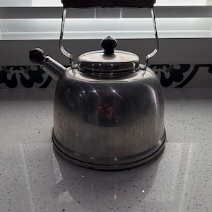 Puede incluir: Una tetera de acero inoxidable para estufa con un asa de madera y un pomo negro en la tapa. La tetera tiene un pico y un asa curva. La tetera está sobre una encimera blanca.