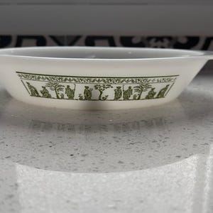 May include: A white, shallow bowl with a green, stylized frieze of figures and trees. The bowl has a slightly flared rim and two small handles. The bowl is sitting on a speckled, light-colored surface.