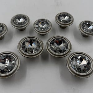 Puede incluir: Un conjunto de diez tiradores de armario plateados con grandes centros de cristal transparente facetado. Los tiradores están dispuestos sobre una superficie blanca, mostrando sus cualidades reflectantes y su diseño decorativo. Ideales para muebles.