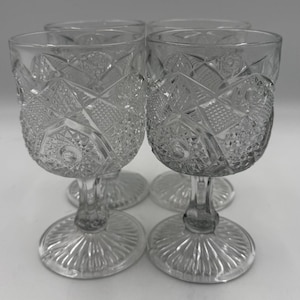 May include: A set of four clear wine glasses with a vintage design. Each glass has a detailed, cut-glass pattern on the bowl and a stemmed base. The glasses are designed with a classic aesthetic, suitable for serving wine or other beverages.