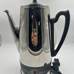May include: A vintage General Electric percolator with a shiny, silver finish. It features a black handle, lid, and base. The coffee pot has a spout for pouring and a power cord with a plug.