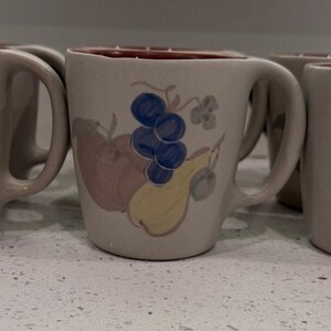 May include: Four beige ceramic mugs with a fruit design. Each mug features a painted illustration of a pear, apple, and grapes in shades of yellow, red, and blue. The interior of the mugs is a dark red color.
