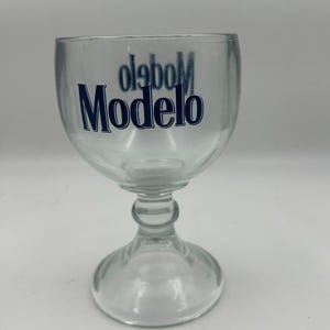Pode incluir: Copo de cerveja Modelo de vidro transparente com uma taça arredondada e uma haste. O logótipo Modelo é impresso em azul na frente do copo. O copo foi concebido para cerveja.