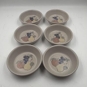 May include: Six small, round, beige ceramic bowls. Each bowl features a painted design of grapes, pears, and apples in shades of blue, yellow, and red. The bowls are arranged in two rows of three.