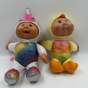 May include: Two Cabbage Patch Kids dolls. One doll is dressed as a unicorn with a white hood, pink bow, and rainbow sequin bodice. The other doll is dressed as a chick with a yellow hood and orange feet. Both dolls have rosy cheeks and green eyes.