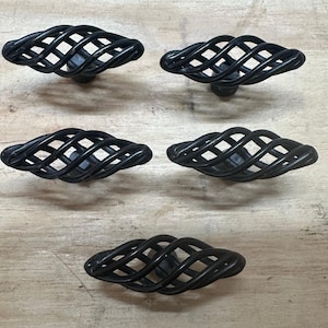 May include: Five black metal cabinet knobs with a cage-like design. The knobs are oval-shaped with a woven pattern, and are set against a light wood grain surface. These decorative knobs are suitable for furniture.