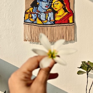 May include: A woven wall hanging depicting a couple in blue, yellow, and red. The artwork is framed by a tan border and fringe. A white flower is held in front of the wall hanging.