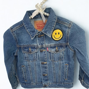 Peut inclure: Veste en jean bleue avec un écusson smiley jaune sur la manche supérieure gauche. La veste a un col classique, des boutons et deux poches de poitrine. Une étiquette rouge Levi Strauss est visible.