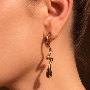 Peut inclure: Une longue boucle d'oreille dorée avec une forme ondulée, lisse et organique qui pend verticalement depuis le lobe de l'oreille. La surface métallique est polie et réfléchissante, avec une forme en goutte au bas de la pièce.