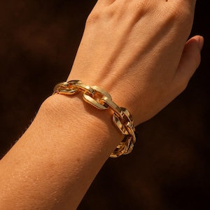 Peut inclure: Un bracelet à maillons de chaîne dorés et épais, composé de grands anneaux ovales polis et entrelacés. Le bracelet présente une finition métallique très brillante et est fixé autour du poignet, affichant un design audacieux et massif.