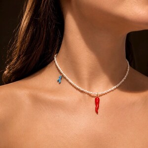 Peut inclure: Un collier de perles blanches composé de deux petits pendentifs : un piment rouge vif suspendu au centre et une petite breloque bleu clair en forme de branche sur le côté, le tout monté sur un délicat cordon de perles blanches arrondies.
