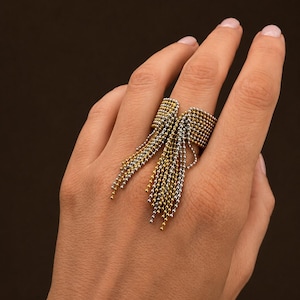 Peut inclure: Une bague composée d'une large bande faite de petites perles métalliques interconnectées dans des tons alternés d'or et d'argent. Un groupe de fines chaînes à billes métalliques pendantes, assorties en or et argent, part du centre de la bague.