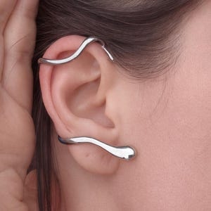 May include: Silver ear cuff jewelry. The ear cuff has a flowing, organic design that wraps around the ear. The jewelry is made of a shiny, metallic material. The ear cuff is worn on the ear and is a modern, minimalist accessory.