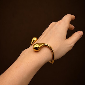 May include: A gold-toned open cuff bracelet featuring two smooth, rounded, teardrop-shaped ends. The metal has a polished, reflective surface that catches the light, creating a sleek and continuous curved design around the wrist area.