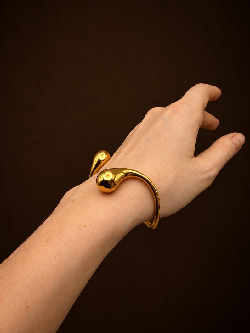 May include: A gold-toned open cuff bracelet featuring two smooth, rounded, teardrop-shaped ends. The metal has a polished, reflective surface that catches the light, creating a sleek and continuous curved design around the wrist area.