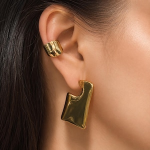 Peut inclure: Gros plan sur une oreille ornée de bijoux en or. L'oreille est ornée d'un ear cuff en or et d'une grande boucle d'oreille en or de forme abstraite. Les bijoux ont une surface polie et réfléchissante, captant la lumière.