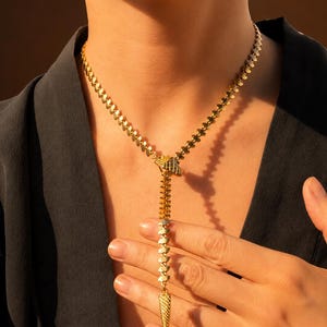 Könnte beinhalten: Eine goldfarbene Halskette mit einer Kette aus kleinen, sich wiederholenden blattförmigen Gliedern. Das Design umfasst ein zentrales Y-förmiges Element, das an einen Reißverschluss erinnert und mit einem strukturierten, kegelförmigen Anhänger endet.