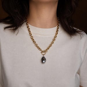 Peut inclure: Un collier à chaîne dorée doté d'un fermoir à bascule sur le devant. Un pendentif unique, irrégulier, gris foncé et à l'aspect métallique est suspendu au centre de la barre de fermeture, créant un contraste marqué avec les maillons dorés.