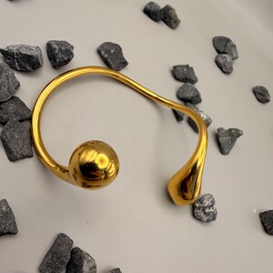 May include: A gold-toned open cuff bracelet featuring a smooth, polished finish. The bracelet has an asymmetrical design with a large, spherical ball at one end and a smaller, teardrop-shaped element at the opposite end, connected by a curved band.