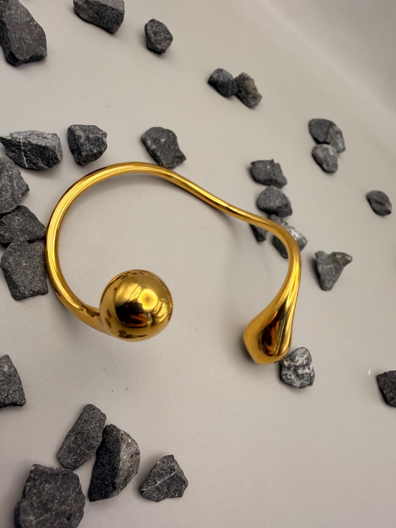 May include: A gold-toned open cuff bracelet featuring a smooth, polished finish. The bracelet has an asymmetrical design with a large, spherical ball at one end and a smaller, teardrop-shaped element at the opposite end, connected by a curved band.