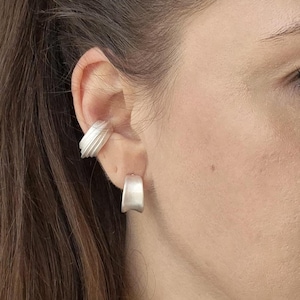 Peut inclure: Gros plan sur une oreille ornée de deux boucles d'oreilles argentées. L'une est une créole incurvée, l'autre une manchette d'oreille texturée. Les bijoux ont une finition mate, reflétant subtilement la lumière. Design moderne et minimaliste.
