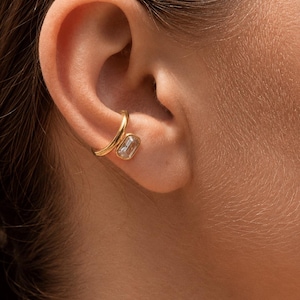 Peut inclure: Une bague d'oreille dorée composée d'un anneau lisse et poli qui entoure le cartilage. Le bijou présente une pierre transparente unique de taille rectangulaire, sertie dans une monture dorée, placée contre la structure de l'oreille.