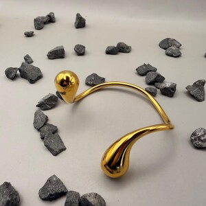 May include: A gold-toned metal collar necklace featuring a curved, open-ended design with two polished, teardrop-shaped terminals at each end. The surface of the metal is highly reflective and smooth, creating a bright, metallic finish.