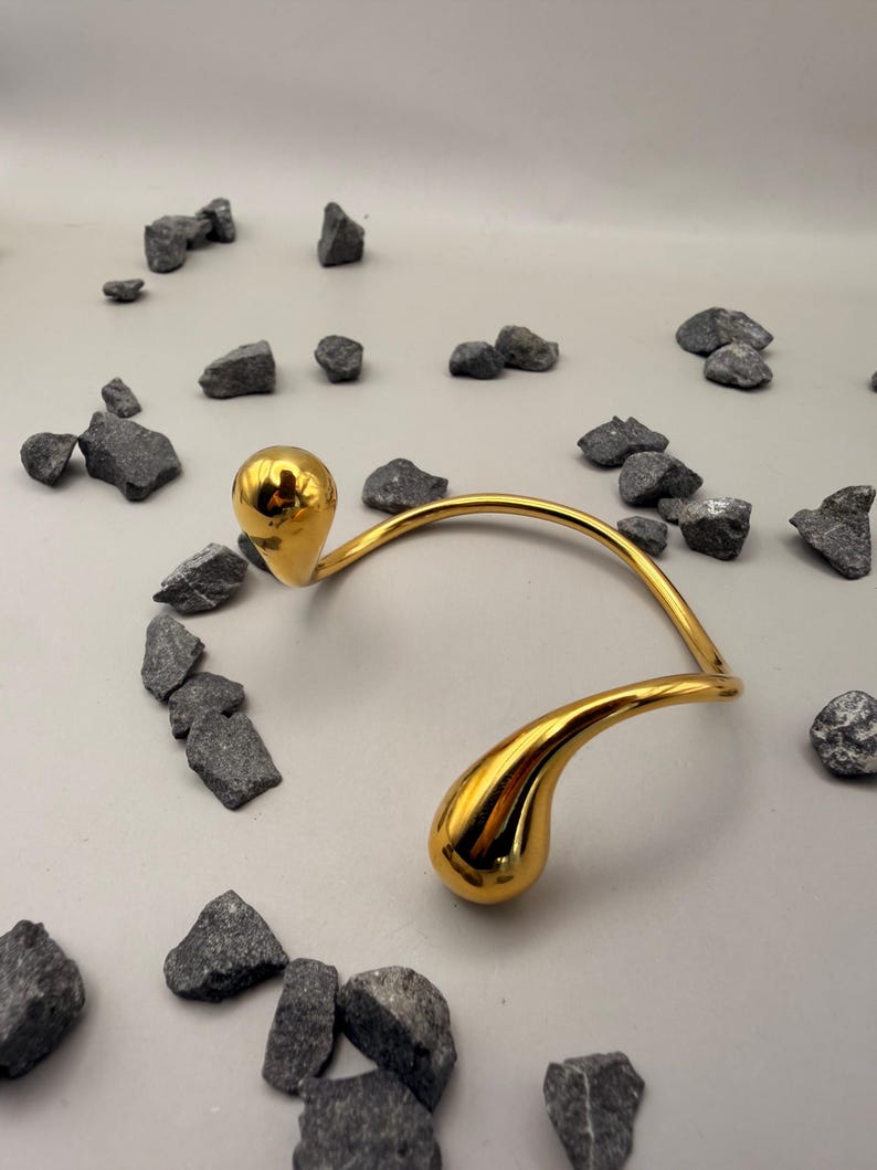 May include: A gold-toned metal collar necklace featuring a curved, open-ended design with two polished, teardrop-shaped terminals at each end. The surface of the metal is highly reflective and smooth, creating a bright, metallic finish.