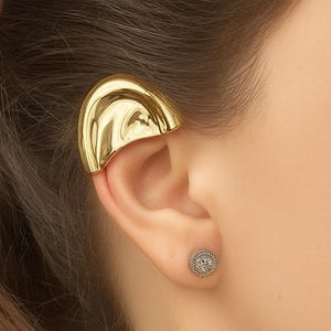 May include: A gold-coloured ear cuff and a small, round stud earring with a halo of small stones are shown. The ear cuff curves over the top of the ear. The earring is in the earlobe.