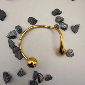 May include: A gold-toned open cuff bracelet featuring a smooth, polished finish. The bracelet has a curved, semi-circular band that terminates in two distinct ends: one side features a spherical ball, while the other side features a teardrop shape.