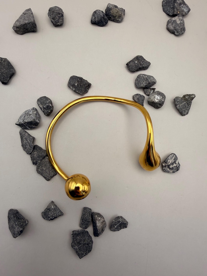 May include: A gold-toned open cuff bracelet featuring a smooth, polished finish. The bracelet has a curved, semi-circular band that terminates in two distinct ends: one side features a spherical ball, while the other side features a teardrop shape.