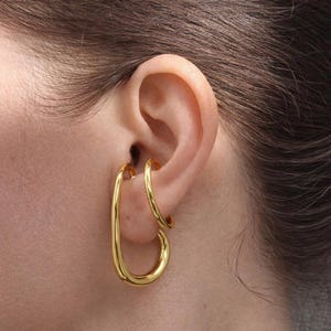 May include: A close-up of a person's ear with two gold earrings. One earring is a small hoop, and the other is a larger, elongated oval shape. The earrings are shiny and reflect light, creating a warm, golden glow.