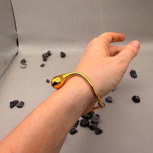 May include: A polished gold-tone metal bangle bracelet featuring a smooth, continuous wave-like design that terminates in a single, rounded, teardrop-shaped end piece. The bracelet has a reflective, high-shine finish and a curved, open-ended structure.