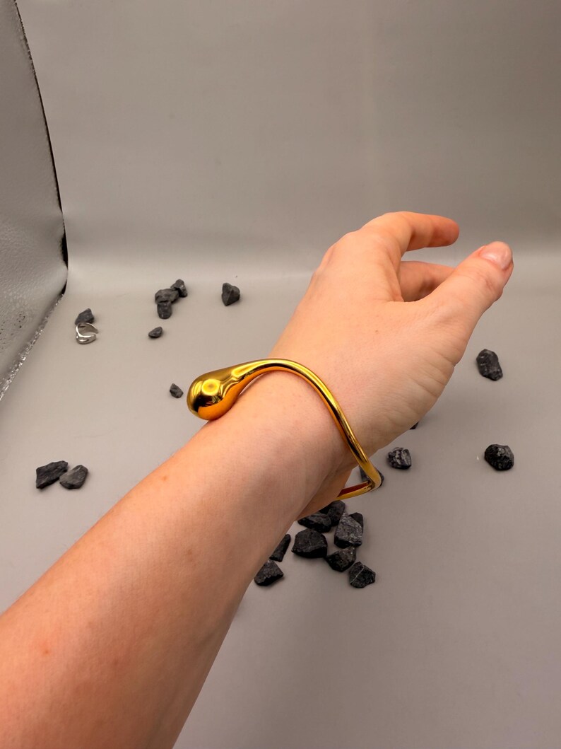 May include: A polished gold-tone metal bangle bracelet featuring a smooth, continuous wave-like design that terminates in a single, rounded, teardrop-shaped end piece. The bracelet has a reflective, high-shine finish and a curved, open-ended structure.