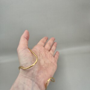 May include: A gold-colored cuff bracelet with a hammered texture. The bracelet has an open design, with a curved shape that wraps around the wrist and forearm. The metal has a slightly irregular surface, giving it a handmade appearance.