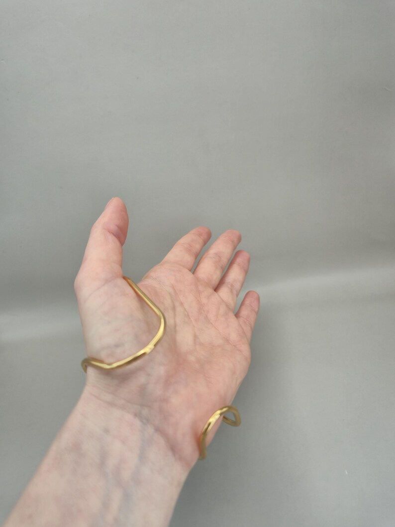 May include: A gold-colored cuff bracelet with a hammered texture. The bracelet has an open design, with a curved shape that wraps around the wrist and forearm. The metal has a slightly irregular surface, giving it a handmade appearance.
