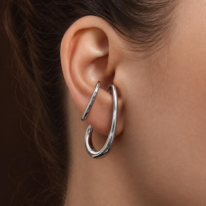 Könnte beinhalten: Ein Paar silberfarbene Ohrklemmen. Eine Manschette hat eine gebogene, längliche ovale Form, die andere ist ein gerader Stab. Der Schmuck wird am Ohr getragen, wobei sich die ovale Manschette um den Rand des Ohrs legt.