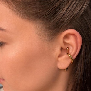 Pode incluir: Um ear cuff de tom dourado com um acabamento liso e polido. A peça de joalharia tem um design curvo de aro aberto que envolve a concha e a área da cartilagem da orelha, com uma pequena protuberância arredondada virada para cima.