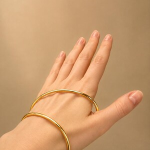May include: Two gold-colored, minimalist bangles. The bracelets are circular and have a smooth, polished finish. The jewelry is displayed on a hand against a neutral background. The bangles are a simple, elegant accessory.