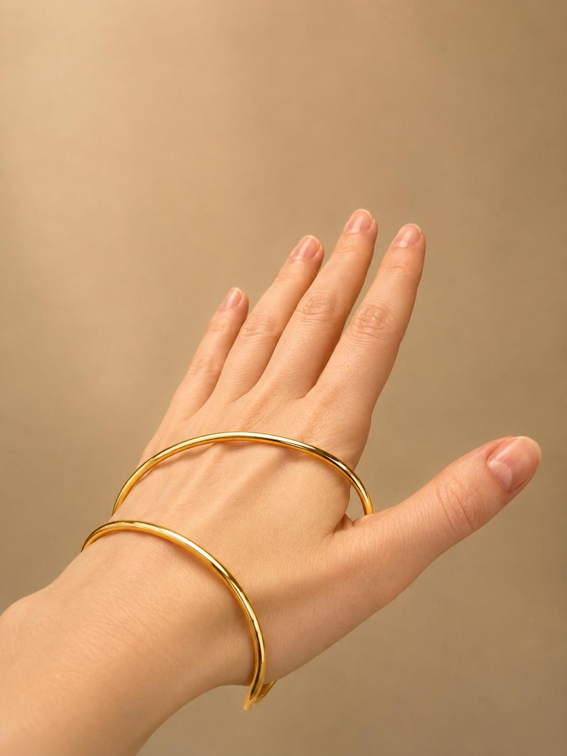 May include: Two gold-colored, minimalist bangles. The bracelets are circular and have a smooth, polished finish. The jewelry is displayed on a hand against a neutral background. The bangles are a simple, elegant accessory.