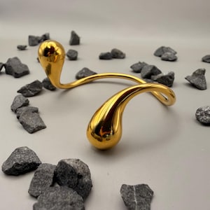 May include: A gold-toned metal collar necklace featuring a smooth, curved wire design that terminates in two polished, teardrop-shaped ends. The piece has a reflective, high-gloss finish and a continuous, open-ended structure that curves gracefully.