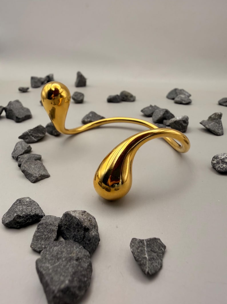 May include: A gold-toned metal collar necklace featuring a smooth, curved wire design that terminates in two polished, teardrop-shaped ends. The piece has a reflective, high-gloss finish and a continuous, open-ended structure that curves gracefully.