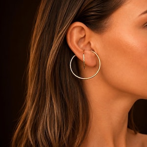 Peut inclure: Une boucle d'oreille créole dorée composée d'un fil métallique fin et circulaire qui passe par le lobe de l'oreille. Le bijou présente un grand anneau extérieur et un segment d'anneau intérieur plus petit, offrant un aspect superposé et lisse.