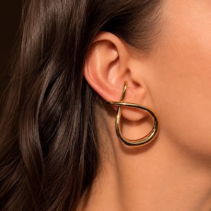 May include: A gold-toned ear cuff featuring a smooth, polished finish and a continuous, fluid loop design that wraps around the outer cartilage of the ear. The metal has a reflective surface and a rounded, sculptural shape that creates a singular, open-ended silhouette.