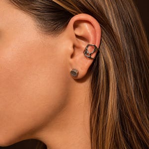 Op de afbeelding: Een zilverkleurige ear cuff met een onregelmatige, organische vorm en meerdere uitsparingen, gedragen op het bovenste kraakbeen, gecombineerd met een kleine, ronde zilverkleurige oorsteker met een textuur op de oorlel.
