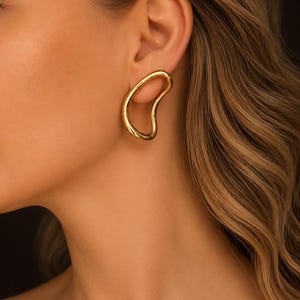 Peut inclure: Une boucle d'oreille dorée présentant une forme irrégulière, organique et ouverte. La boucle d'oreille possède une finition métallique lisse et polie, portée sur le lobe de l'oreille, affichant un design continu, fluide et asymétrique.