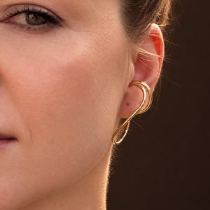 Peut inclure: Une boucle d'oreille en métal doré au design continu à double boucle qui s'enroule autour du bord extérieur de l'oreille. Le métal présente une finition polie et réfléchissante, créant une silhouette incurvée et élégante.