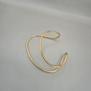 May include: A gold-colored cuff bracelet with a unique, open design. The bracelet features a continuous, flowing shape, creating a modern and minimalist aesthetic. The metal has a polished finish, reflecting light and adding a touch of elegance.